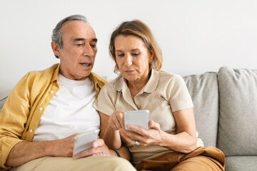 Senior couple sitting on sofa and using smartphone, trying to understand mobile application,...