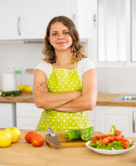 Obraz na płótnie Canvas Positive woman wearing apron, standing at kitchen table while preparing salad.