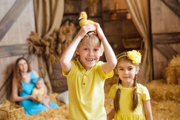 boy holds a yellow duckling on his head, and his sister stands next to him, both laughing. They are standing in front of a rural barn with hay bales. - Powered by Adobe