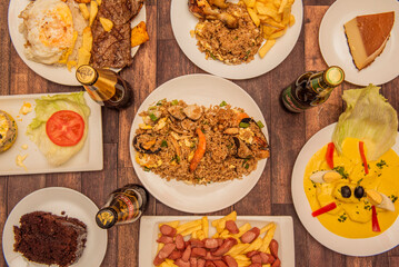 A set of Peruvian food dishes with papas a la huancaina in the center, a quesillo, steaks with rice and plantains, arros chaufa and drinks on a brown table