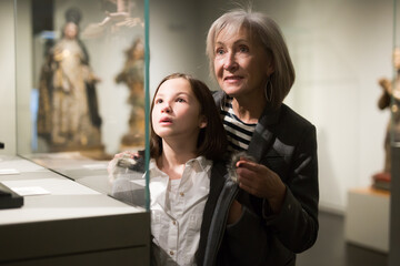 Inquisitive preteen girl visiting museum of ancient sculpture with her grandmother, exploring antique showpieces together