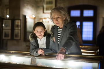 Friendly aged female tutor helping preteen girl exploring antique showpieces in local history museum..