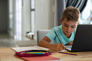 Caucasian boy writing notes in book while doing homework online over laptop at home, copy space