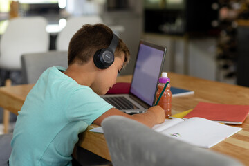 Caucasian boy wearing headphones writing homework by laptop on table at home, copy space