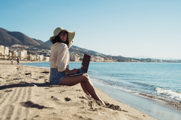 young beautiful girl freelancer with a laptop works on the seashore sitting on the sand