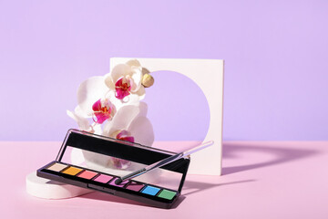 Palette of eyeshadows, brush, orchid flowers and plaster figure on lilac background