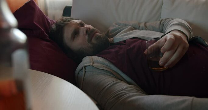 Depressed alcoholic man with an alcohol addiction problem holding a whiskey glass in hand. Feel bad lying on the couch. Make decision to give up alcohol. Binge drinking. Alcohol rejection concept 