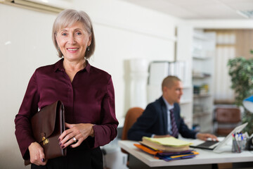 Portrait of smiling elegant elderly business woman standing in office holding briefcase of documents