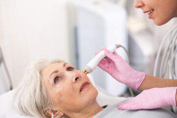 Obraz na płótnie Canvas Delighted old woman lying on clinical chair in aesthetic cabinet during face RF-lifting procedure