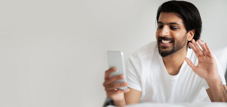 Happy young arabic male in white t-shirt, waving hand, has video call on smartphone in room