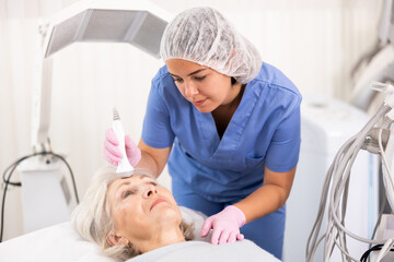 Delighted old woman lying on clinical chair in aesthetic cabinet during face laser therapy procedure
