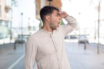 young handsome man looking bewildered and astonished, with hand over forehead looking far away, watching or searching