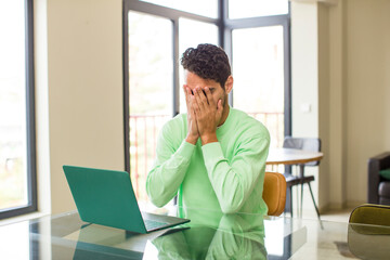 young hispanic man feeling sad, frustrated, nervous and depressed, covering face with both hands, crying. working at home