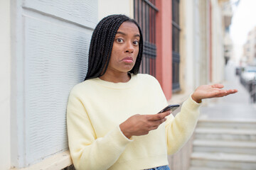 black afro woman shrugging, feeling confused and uncertain. using a smartphone concept