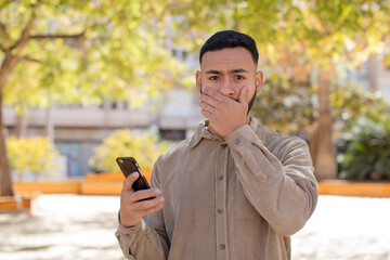 young  adult man covering mouth with a hand and shocked or surprised expression. using a smartphone...