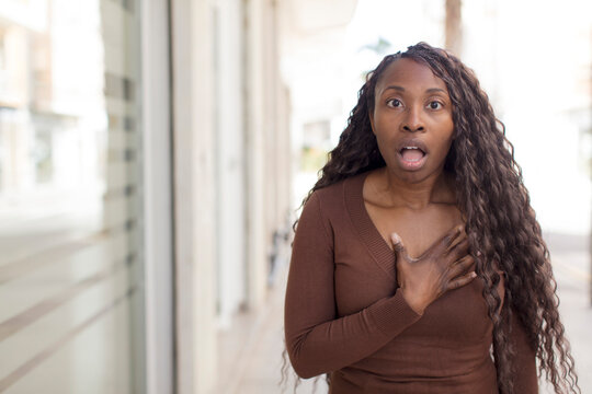 afro pretty black woman feeling shocked, astonished and surprised, with hand on chest and open mouth, saying who, me?