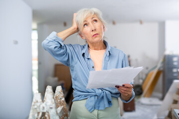 Portrait of frustrated elderly woman interior designer checking documents in apartment during...