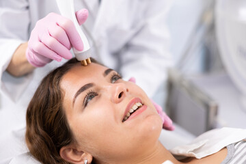 Naklejka premium Elderly beautician doctor doing RF lifting procedure for flawless woman face laying in beauty salon. Hardware cosmetology.