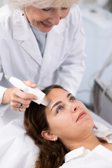 Pleased young woman having laser peeling procedure of face by means of apparatus for aesthetic procedures used by specialist