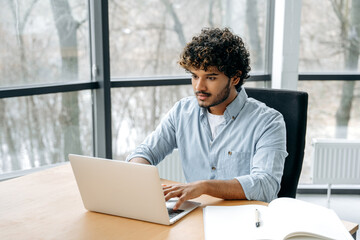Fototapeta premium Handsome successful indian or arabian curly man, financial manager, ceo, IT specialist, sitting at a desk in the modern office, focused looking at a laptop screen, working on a project or with clients