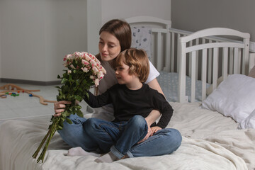 Cute little toddler boy stand hiding flowers bouquet and gift making birthday surprise for young mother, small child son give present congratulate mom on the bed. Mothers day. Happy family.