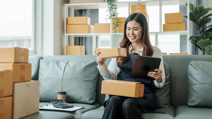 Using tablet computer, Young pretty Asian female influencer small businesses SME owners female entrepreneurs working on receipt box and check online orders, sitting on sofa of home online business.