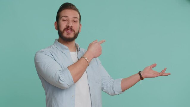 Young lebanese man showing thumbs up and pointing empty place, advertising area for commercial text, copy space for goods promotion. Bearded arabian guy isolated on blue studio background, indoors