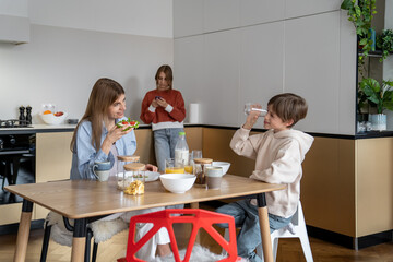 Happy young mother with indulging child son having fun breakfast eat sandwich and cornflakes at home, while teen girl daughter standing with smartphone focused looking at screen on kitchen in morning