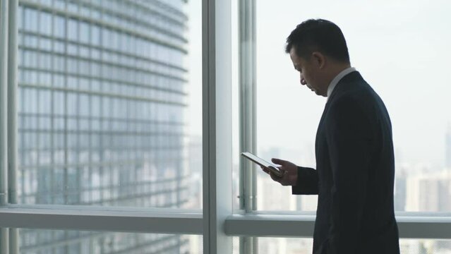asian business man pacing by the window while looking at cellphone
