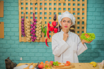 Beautiful asian female professional chef holding a bowl of fresh green vegetables in kitchen full of cooking ingredients with doubtful expression.