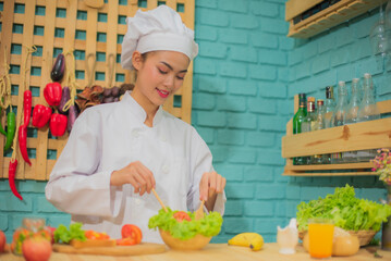 Beautiful asian female professional chef stirring bowl of fresh green vegetables with a spatula in kitchen full of cooking ingredients with smile on her face.