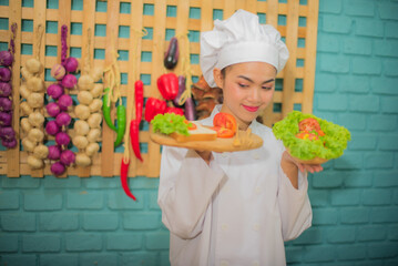 Side view of beautiful asian female professional chef holding and looking to a wooden tray with tomatoes bread and bowl of fresh green vegetables in kitchen.