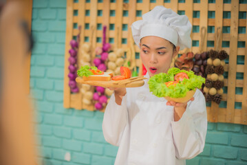 Side view of beautiful asian female professional chef holding and looking a wooden tray with tomatoes bread and bowl of fresh green vegetables in kitchen with joyful expression.