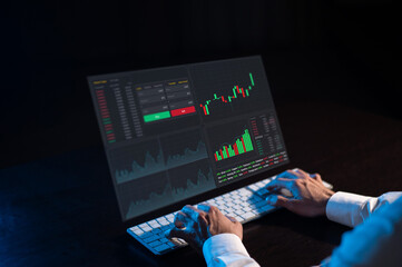 Caucasian man sits in the dark in front of a virtual screen. Stock exchange charts. Close-up of male hands on the keyboard.