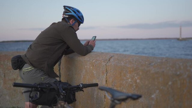 Cycling trip to sea at sunset. Male cyclist is resting on embankment by sea and uses smartphone to build route and navigate GPS. Male with bike on seashore surfing internet on a smart phone.