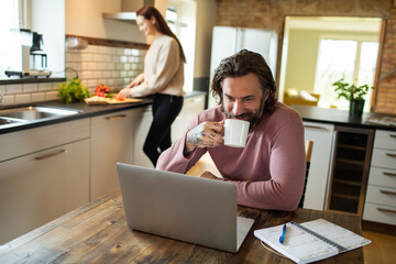 Young adult man using a laptop while his wife is working in the kitchen behind him