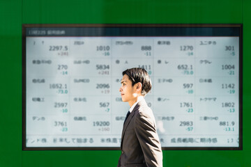 屋外の電光掲示板に映る株価ボードの前を歩いて通り過ぎる30代の日本人のスーツ姿のビジネスマン