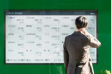 電光掲示板の株価ボードを見てその不景気に首に手を当てて落胆する30代のスーツ姿のビジネスマンの後ろ姿 - obrazy, fototapety, plakaty