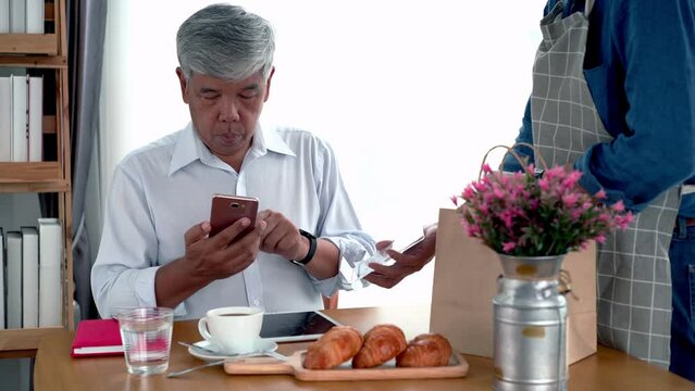 Active senior Asian man working in a coffeeshop using a mobile device, smartwatch for NFC, QR code scanning, contactless payment to pay for coffee and snack. Active Senior concept.