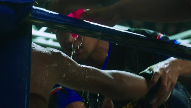4K Cinematic slow motion footage of a Muay Thai coach pouring water on his Thai fighter and massaging his arm on a boxing ring during a Muay Thai boxing fight in a stadium