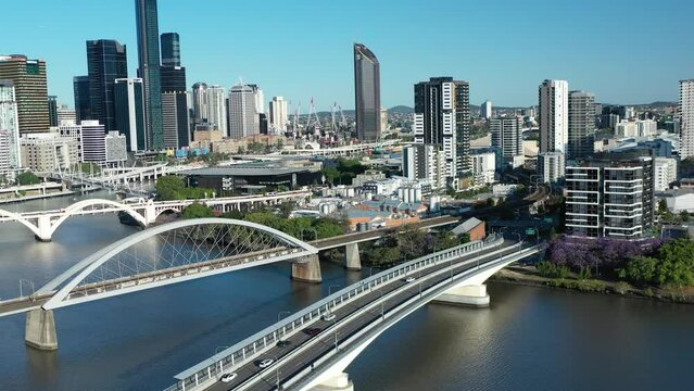 Aerial shot flying backwards of Brisbane Go Between Toll Bridge. Cars and cyclists crossing the bridge in the evening sunlight. City Cat Boat drives underneath bridge. Brisbane City in background