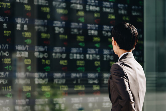 ビジネス街に設置された屋外電子掲示板の株価ボードを見る30代の日本人のビジネスマンの後姿