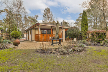 a house in the middle of a garden with trees and shrubs around it there is a picnic table on the lawn