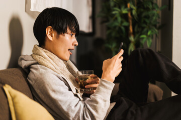自宅の部屋のソファに座ってスマートフォンを見て驚く表情をする30代の日本人男性