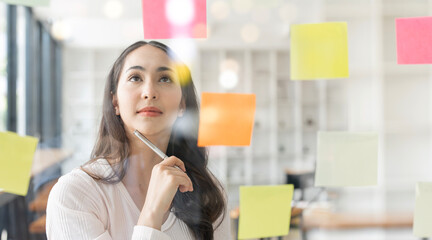 Young smiley attractive businesswoman using sticky notes in glass wall to writing strategy business...
