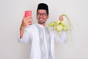 Moslem Asian man smiling happy when calling his family while showing rhobus shaped rice cake