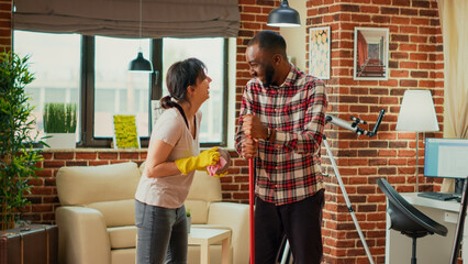 Life partners doing cleaning chores together in apartment, mopping floors and sweeping dust off. Diverse people enjoying spring cleaning session with mop and furniture polish. Handheld shot.