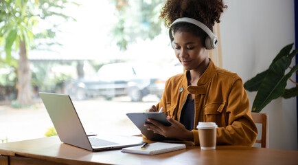 Young African woman listening music from headphones and writing note for her work idea in diary book.She in home office
