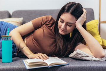 Teen girl lying on the couch and reads a book. Modern teenager at home. Teen literature concept.