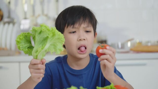 Asian little boy frowning unhappy and don't like eating vegetables holding greens vegetables and tomato on the tabel at kitchen room. Nutrition & healthy eating habits for kids concept